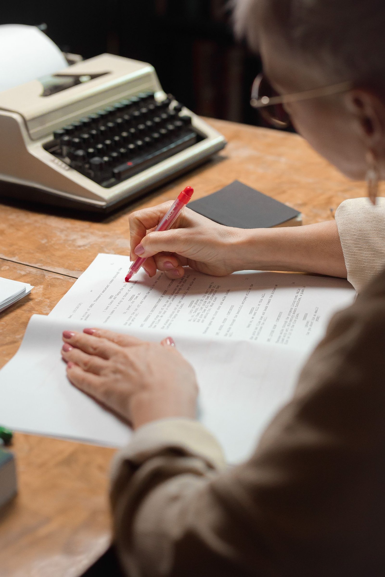 A woman reviews and edits papers using a red pen near a vintage typewriter.